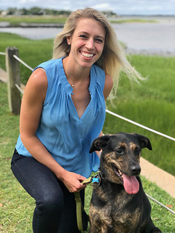 A woman kneeling on grass next to a dog, both smiling at the camera, with a body of water behind them.