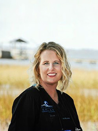 The image shows a woman standing outdoors with water and a building visible behind her. She appears to be wearing professional attire, possibly related to a medical profession, given the stethoscope around her neck.