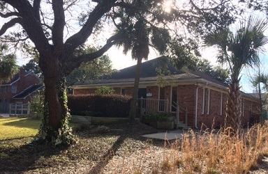 The image depicts a single-story house with a red brick exterior, situated in a residential area with trees and a clear sky.