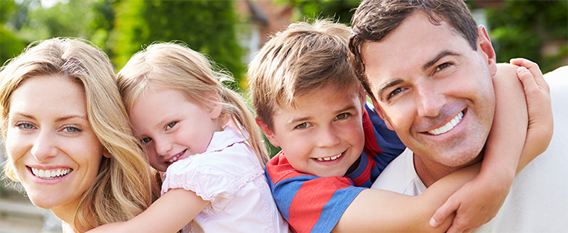The image shows a family of four people, including two adults and two children, embracing and smiling at the camera.
