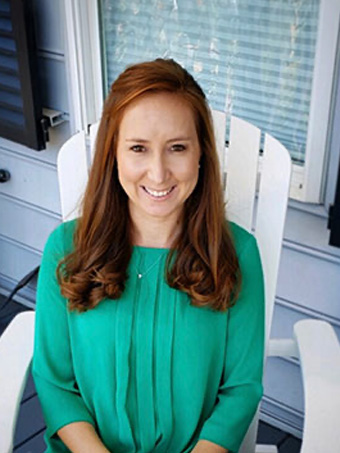 The image shows a woman sitting on a porch, smiling at the camera. She has long brown hair and is wearing a green top with short sleeves.