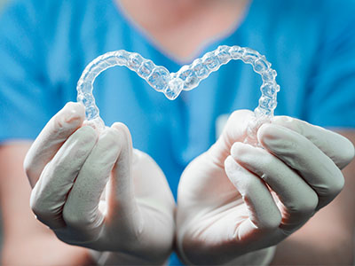 The image shows a pair of transparent dental retainers being held by a person wearing gloves, with a blue background.