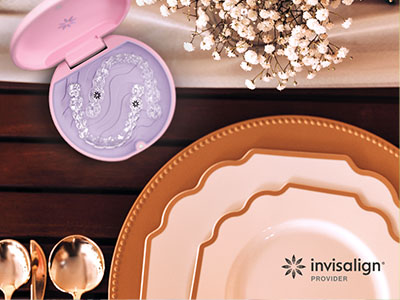 The image displays a collection of tableware items including plates, bowls, and a toothbrush holder, arranged on a wooden surface with a pink lid open, set against a backdrop of a dining table and a floral arrangement.