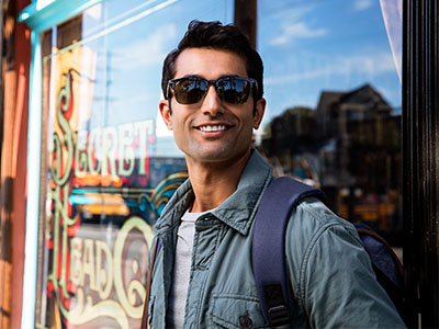 A man wearing sunglasses and a backpack, standing outdoors in front of a storefront with a sign.