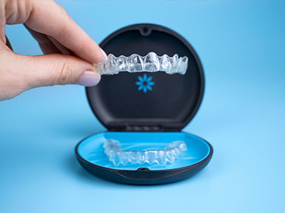 The image shows a hand holding an open case containing a set of clear dental aligners, with one aligner partially inserted into a blue holder.