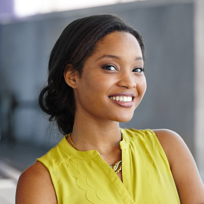 A young woman with dark hair smiling, wearing a yellow top and posing against a blurred background.