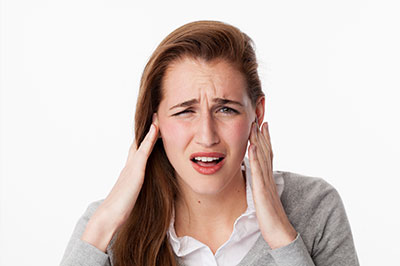 The image shows a woman with her hand on her head, appearing to be experiencing pain or discomfort, possibly due to a loud noise or pressure, as indicated by her facial expression and gesture.