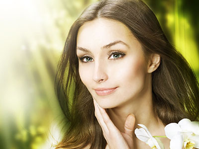 A woman with fair skin, long hair, and makeup looking directly at the camera against a blurred background of greenery.