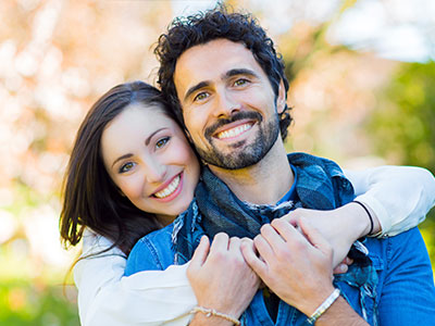 The image shows a man and woman embracing each other with smiles, set against a blurred background.