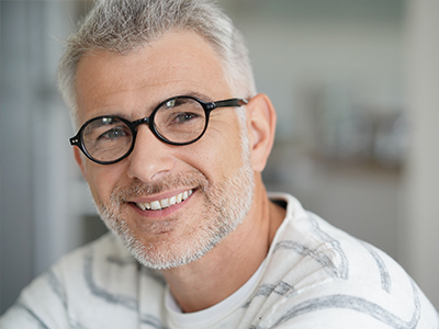 The image depicts a middle-aged man with glasses, wearing a white shirt, smiling at the camera.