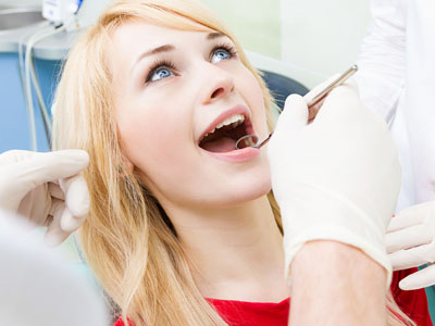 A woman sitting in a dental chair with her mouth open, receiving dental treatment from a dentist who is examining her teeth.