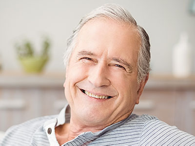 The image shows a smiling older man with gray hair, wearing a blue shirt, sitting comfortably in a living room environment.
